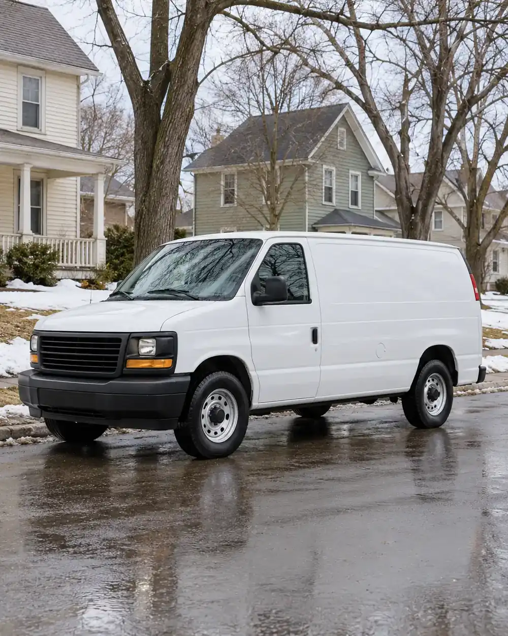 Unmarked white work van on a residential street with mature trees, wet pavement, and melting snow.